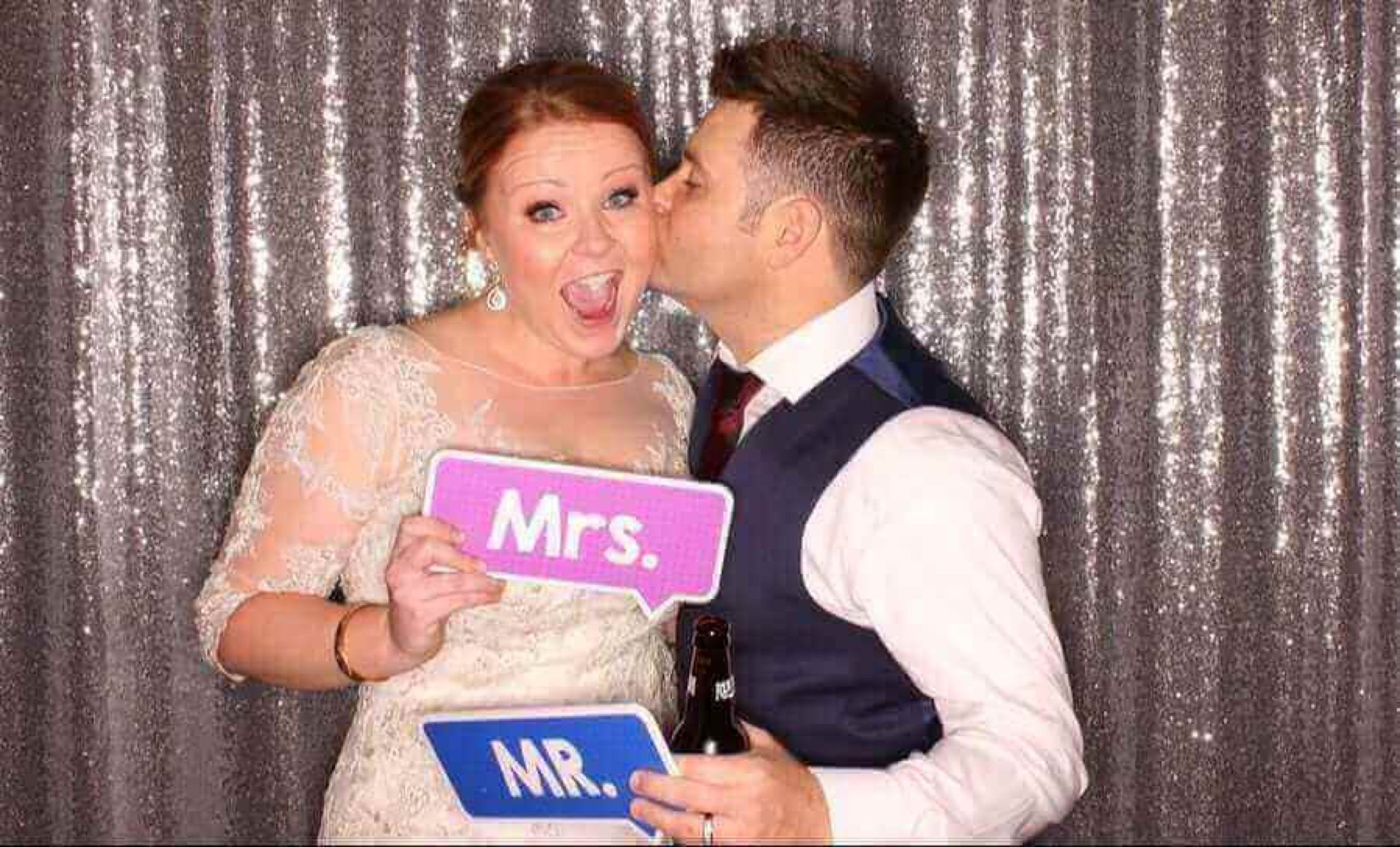 Groom kissing the laughing bride's cheek in a wedding photo booth, the two of them holding Mr. and Mrs. signs against a silver sequin backdrop