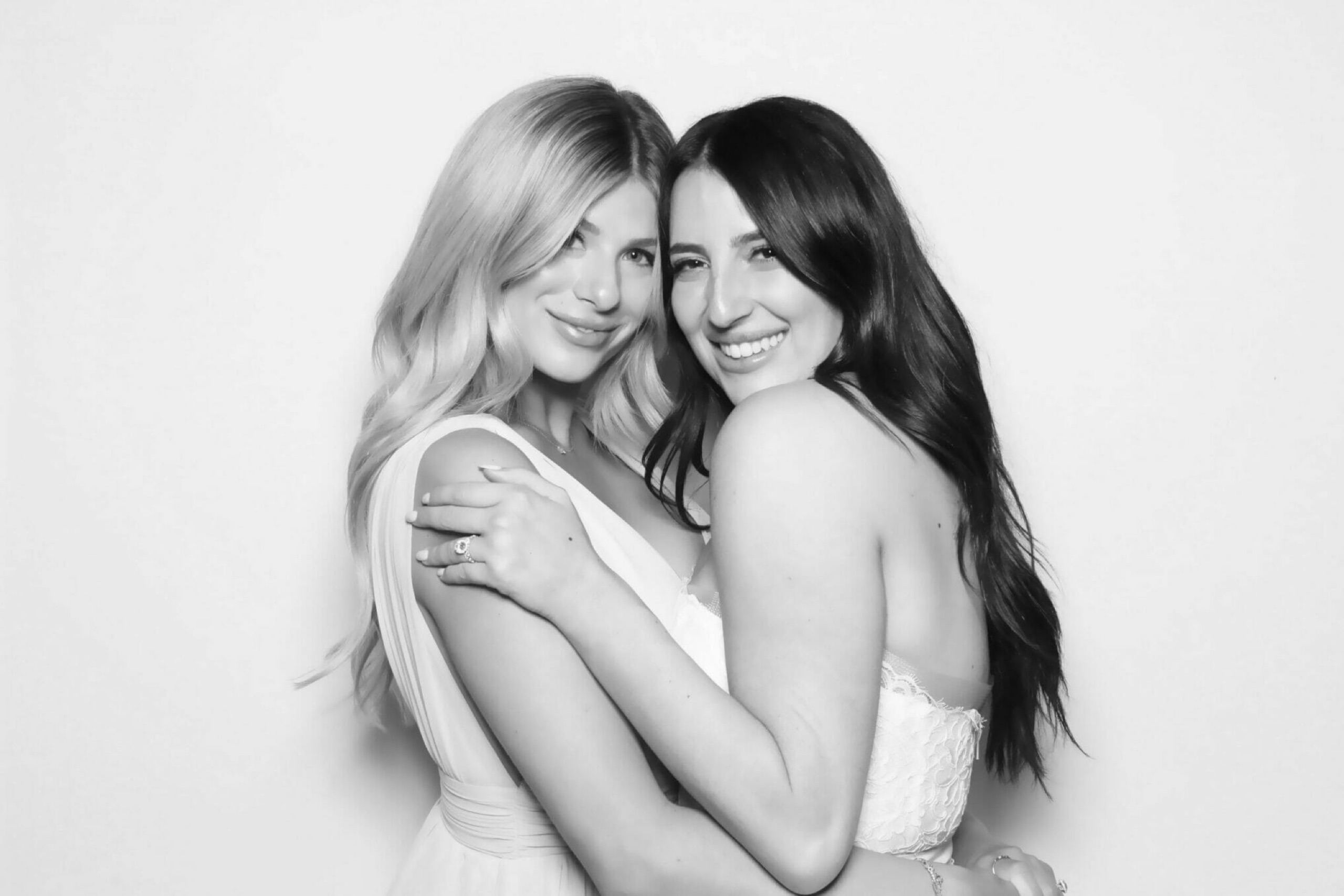 Two smiling guests in white dresses posing cheek to cheek for a black-and-white glam booth portrait against a bright white backdrop