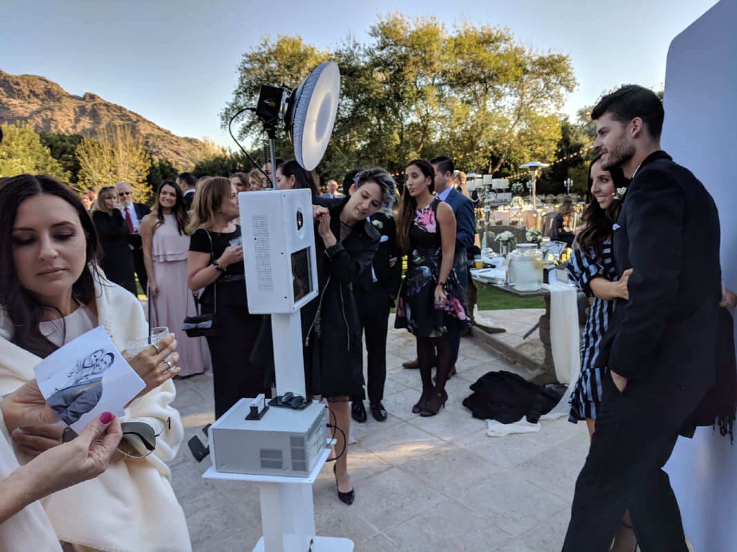 Open-air photo booth setup with studio lighting at an outdoor desert wedding, guests waiting their turn beside the backdrop