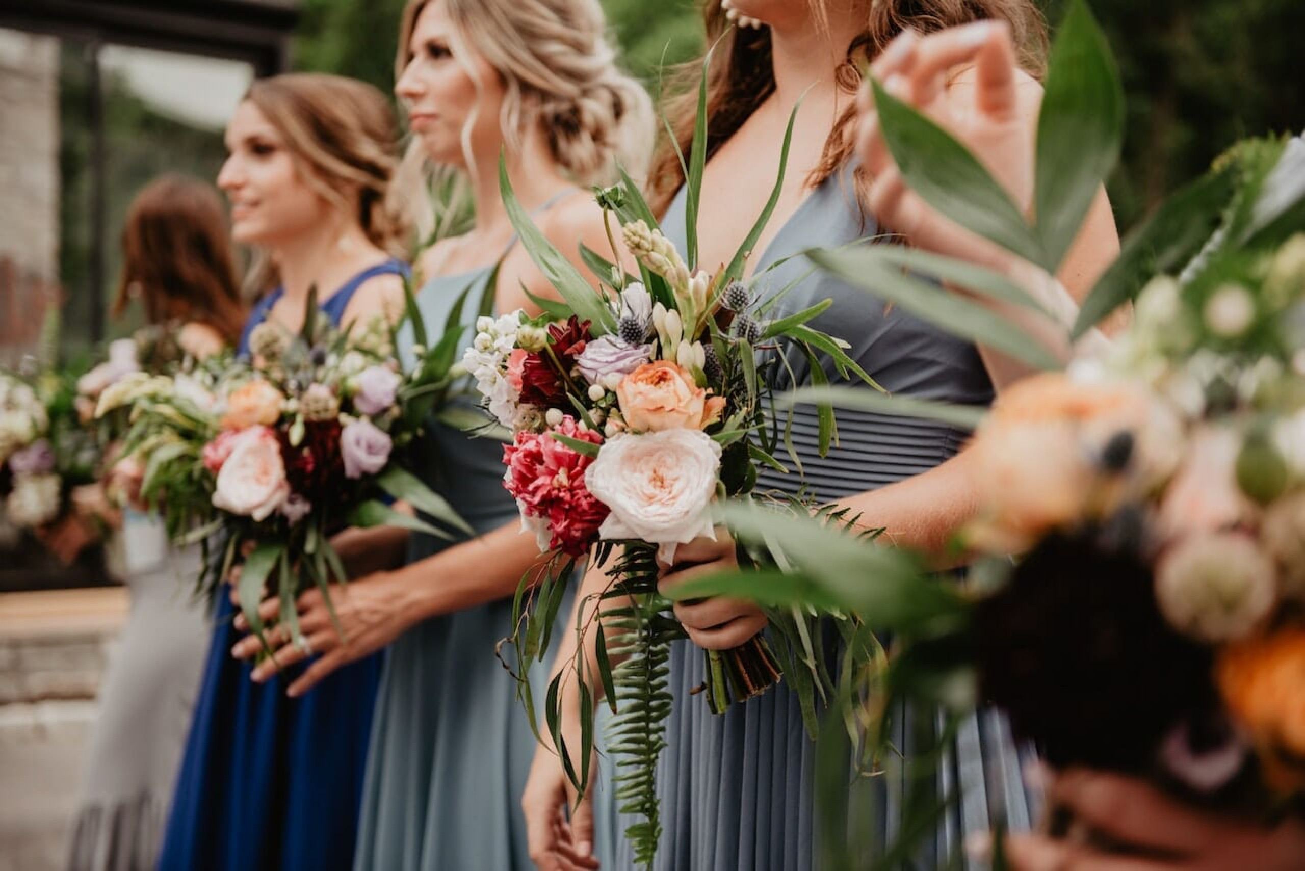 Bridesmaids lined up in mismatched blue and gray dresses holding tropical greenery bouquets