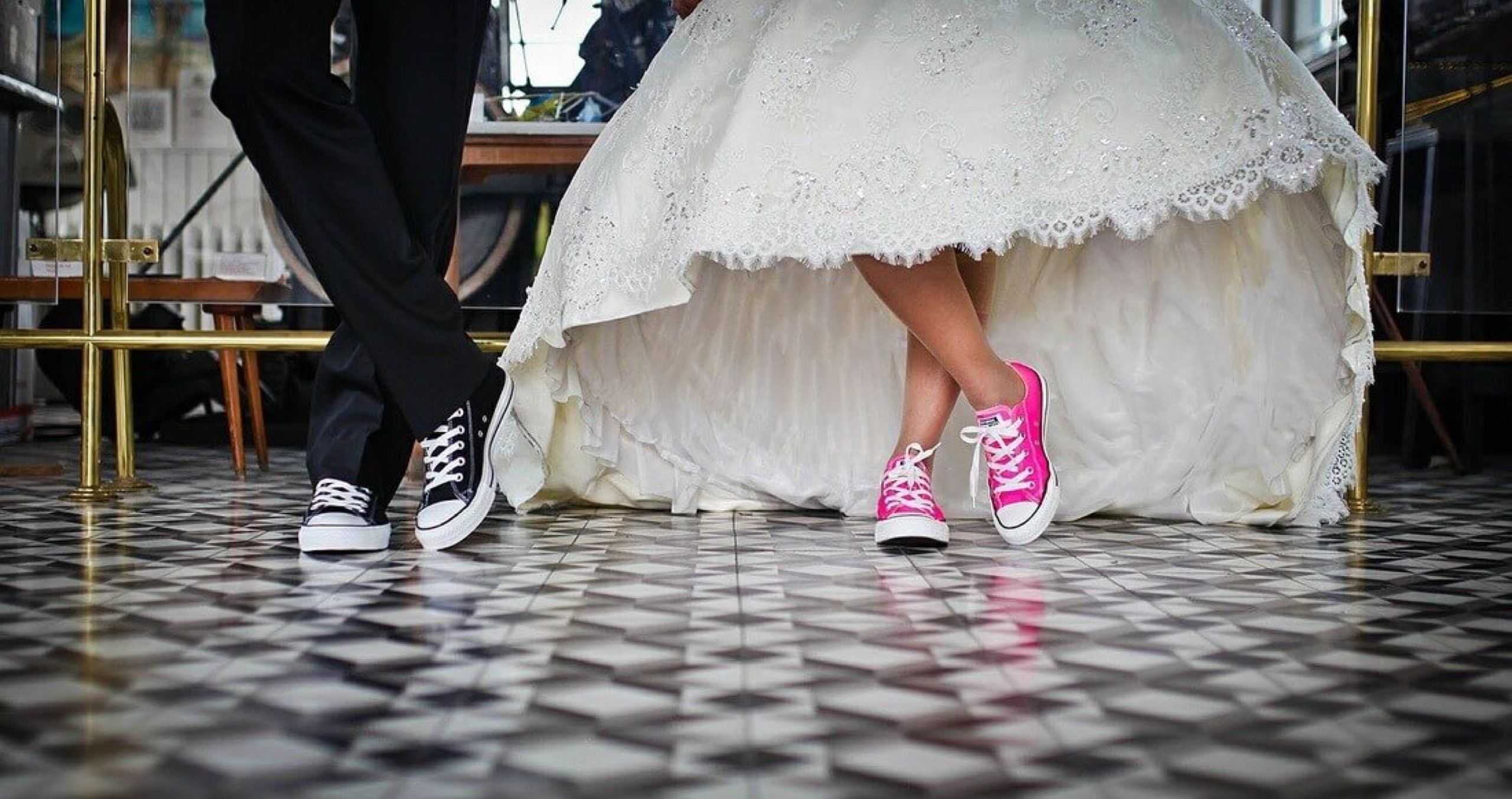 Bride in a lace ballgown wearing hot pink sneakers standing beside a groom in a black tuxedo and black sneakers on a checkered tile floor