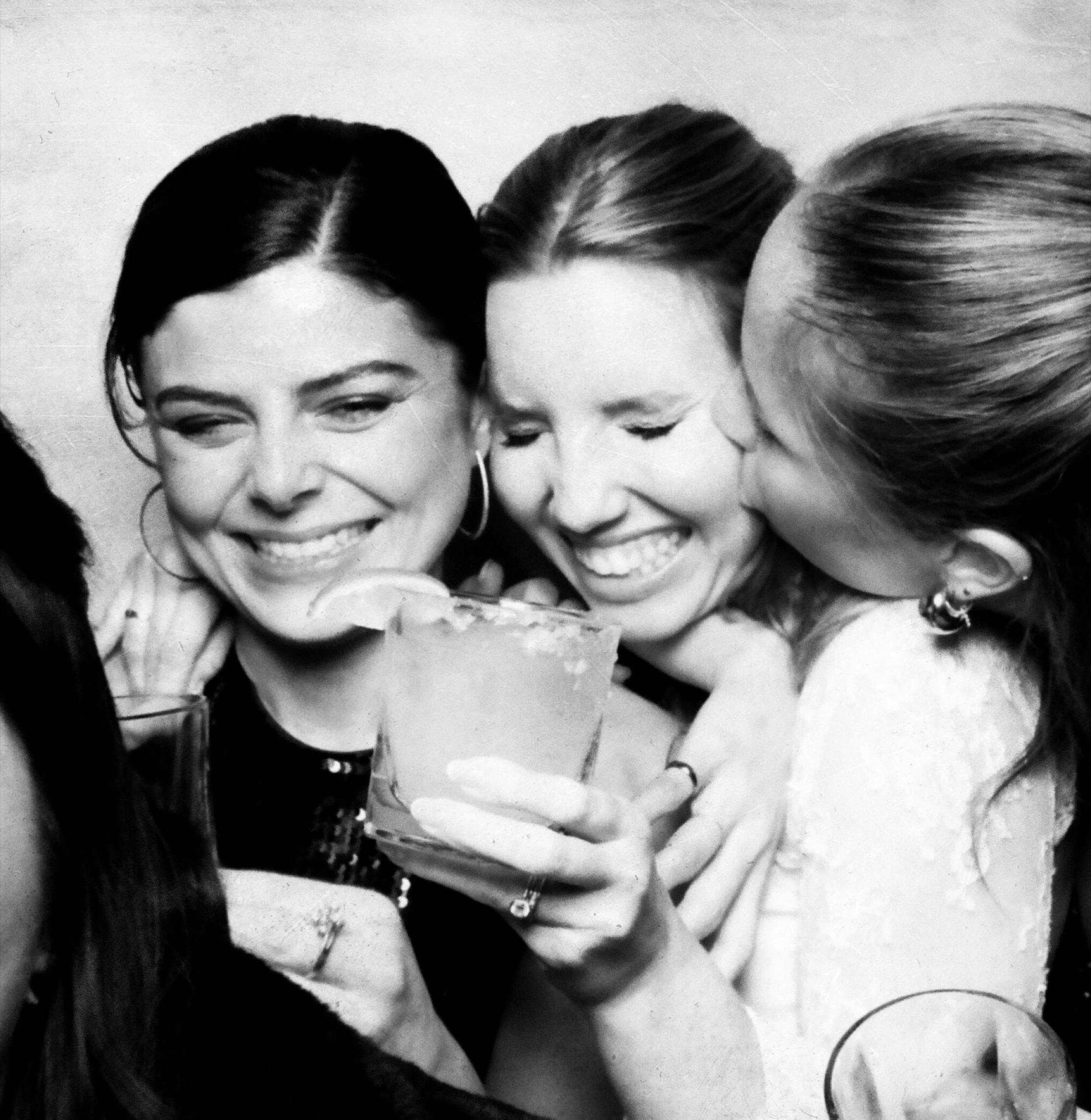 Three guests laughing and hugging, drinks in hand, in a black-and-white vintage photo booth shot at a Valley celebration