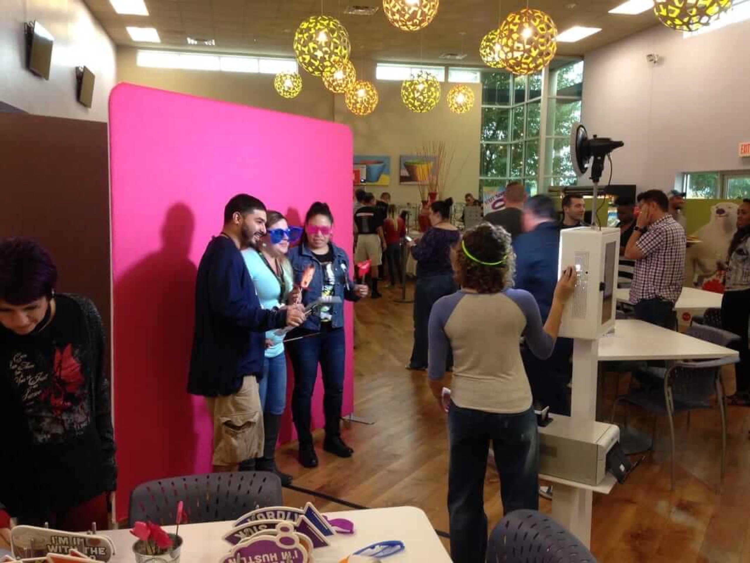 Guests posing with props against a hot pink backdrop while an attendant runs the open-air booth beside a props table