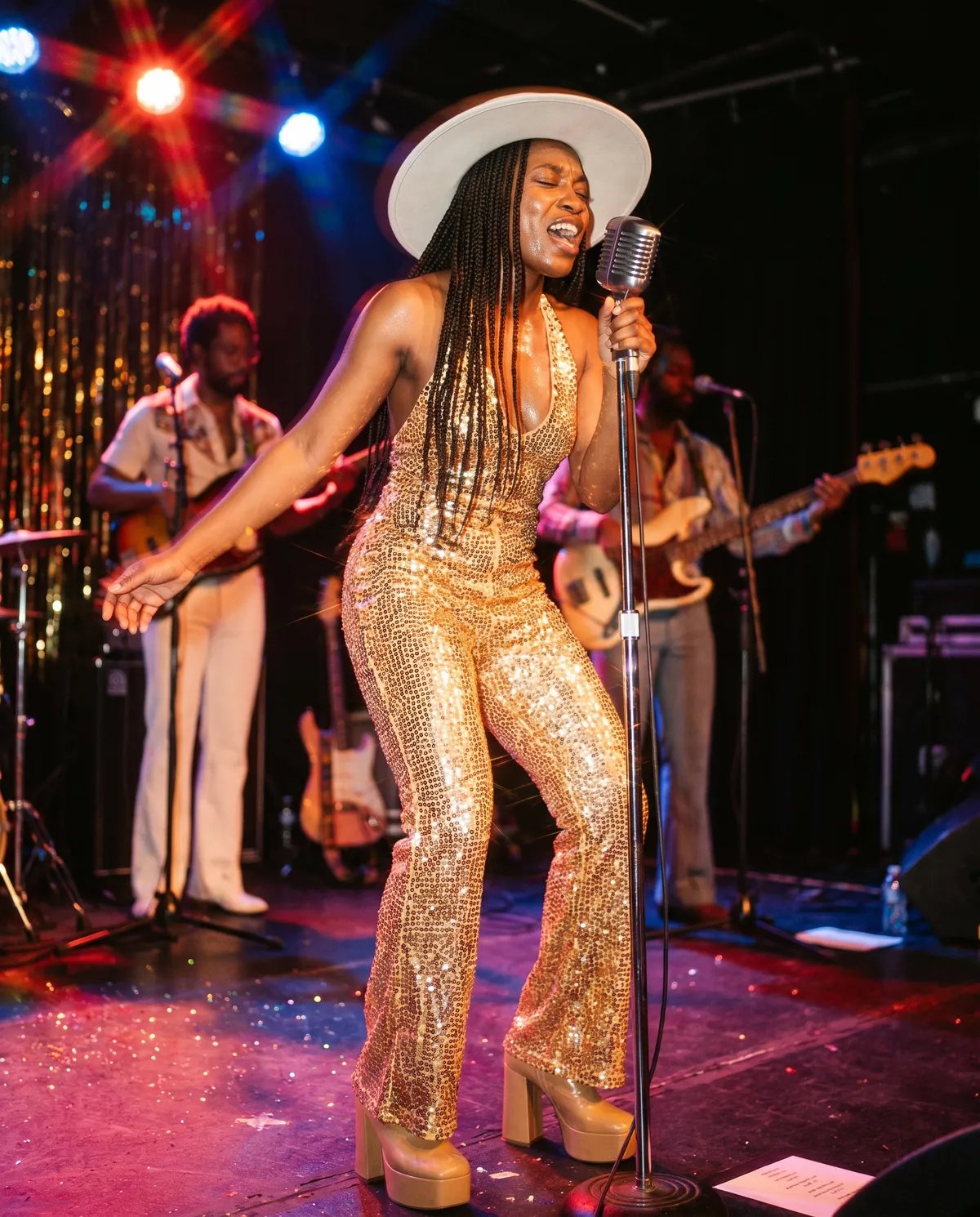 The guest as a 1975 funk singer in a gold sequined jumpsuit belting into a vintage microphone